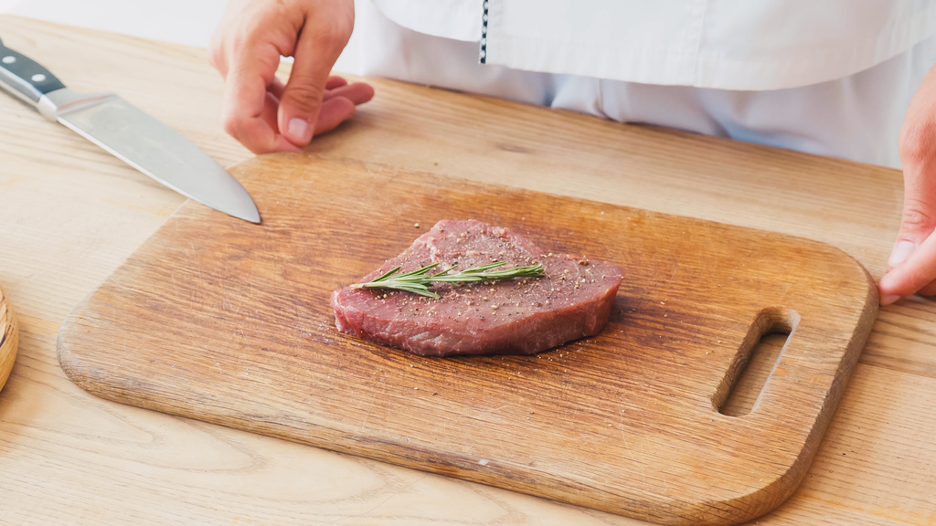 vista parcial del chef de pie cerca de filete de res con rama de romero en la tabla de cortar - Foto, imagen