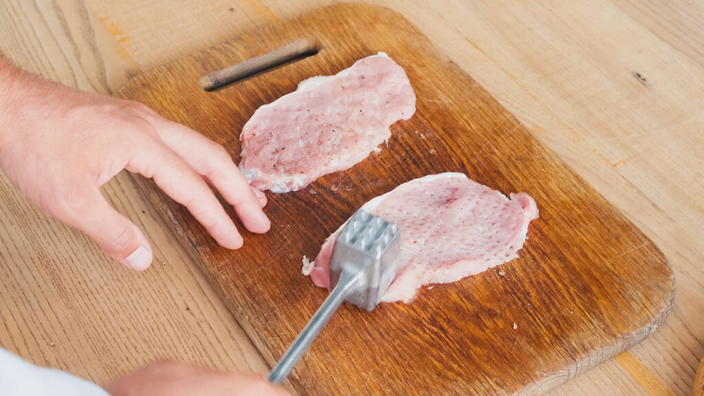 gedeeltelijk zicht van chef-kok met hakhamer afvlakkend varkensvlees op tafel - Foto, afbeelding