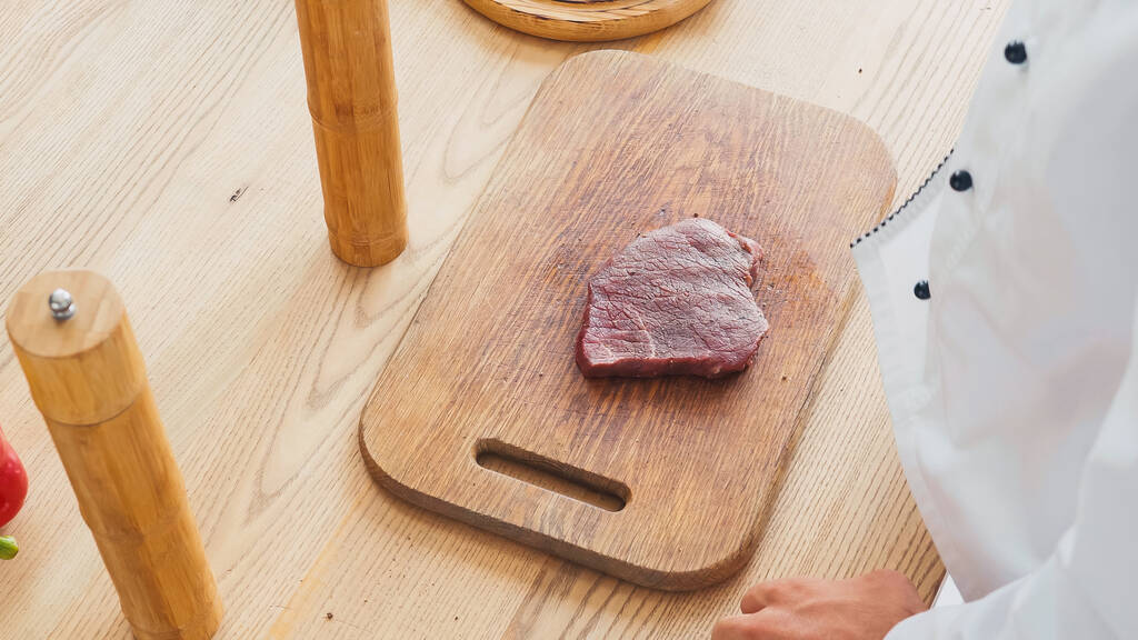 Vista recortada del chef de pie sobre el filete de carne de res en la tabla de cortar - Foto, Imagen