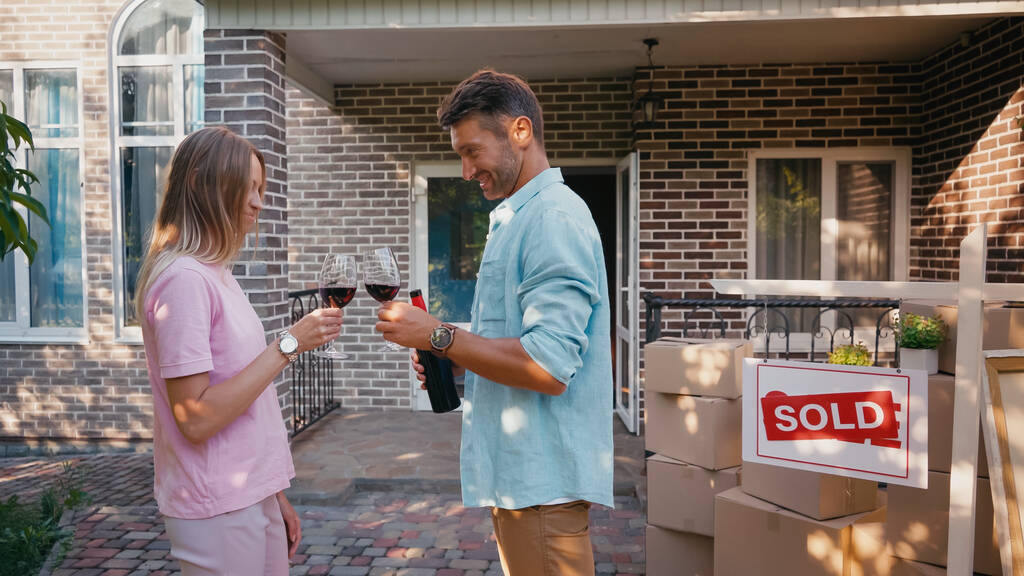 πλαϊνή άποψη του ευτυχισμένου ζευγαριού clinking ποτήρια του κόκκινου κρασιού κοντά στο νέο σπίτι  - Φωτογραφία, εικόνα