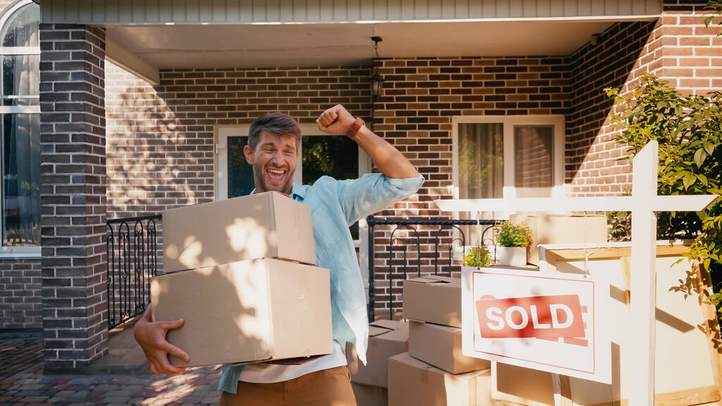 hombre excitado sosteniendo cajas y regocijándose cerca de casa nueva - Foto, imagen
