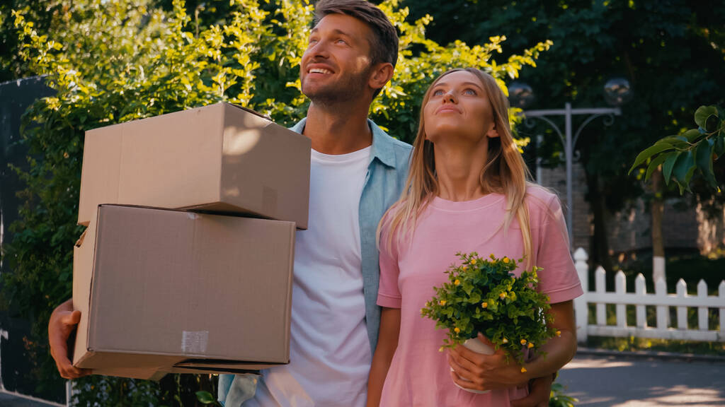alegre esposo y esposa caminando con cajas y plantas, concepto de reubicación  - Foto, Imagen