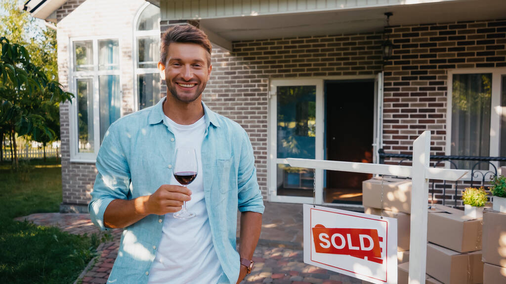 joyful man holding glass of red wine near new house  - Photo, Image
