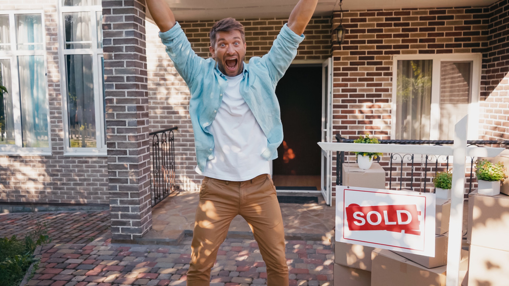 взволнованный мужчина с поднятыми руками и открытым ртом у доски с проданной повязкой и новым домом  - Фото, изображение