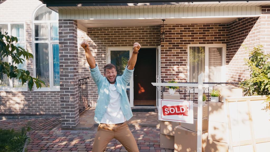радісний чоловік з піднятими руками і відкритим ротом біля дошки з проданим написом і новим будинком
  - Фото, зображення