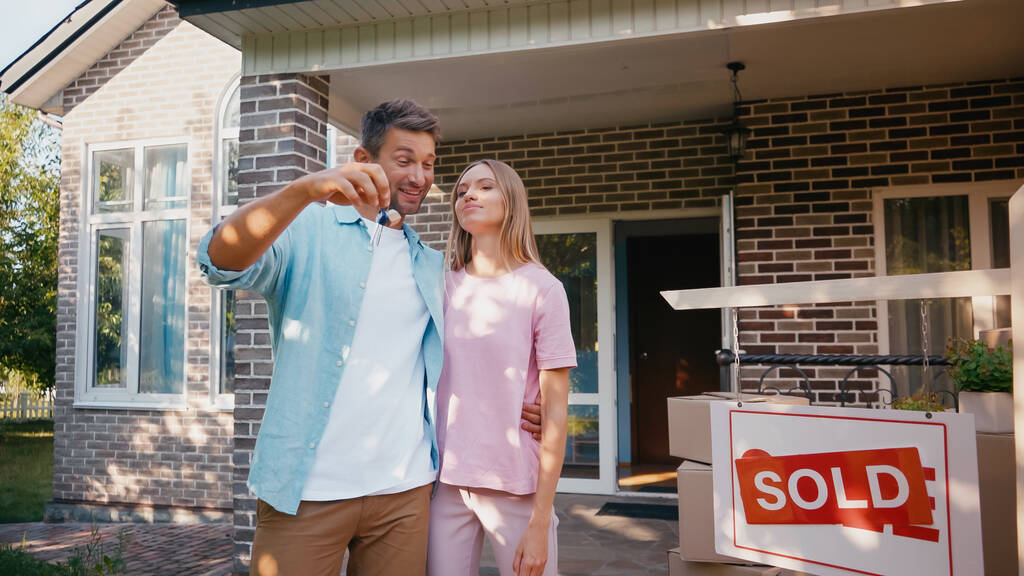ευτυχισμένος άντρας που κρατάει κλειδιά και αγκαλιάζει τη γυναίκα του κοντά στο νέο σπίτι και πουλάει σανίδες  - Φωτογραφία, εικόνα