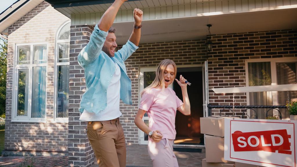 счастливый мужчина держит ключи рядом с женой и танцует возле нового дома и продает доску  - Фото, изображение
