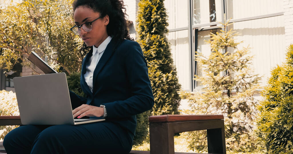 Kıvırcık Afrikalı Amerikalı iş kadını dizüstü bilgisayar kullanıyor ve bankta otururken not defteri tutuyor.  - Fotoğraf, Görsel