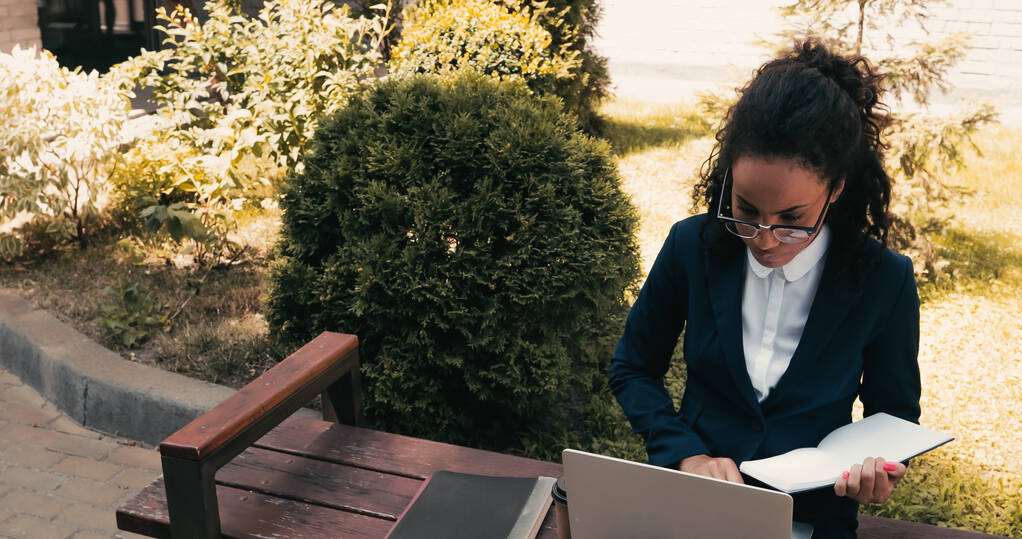 Afrikai amerikai üzletasszony szemüvegben tartja notebook és ül a padon laptop  - Fotó, kép