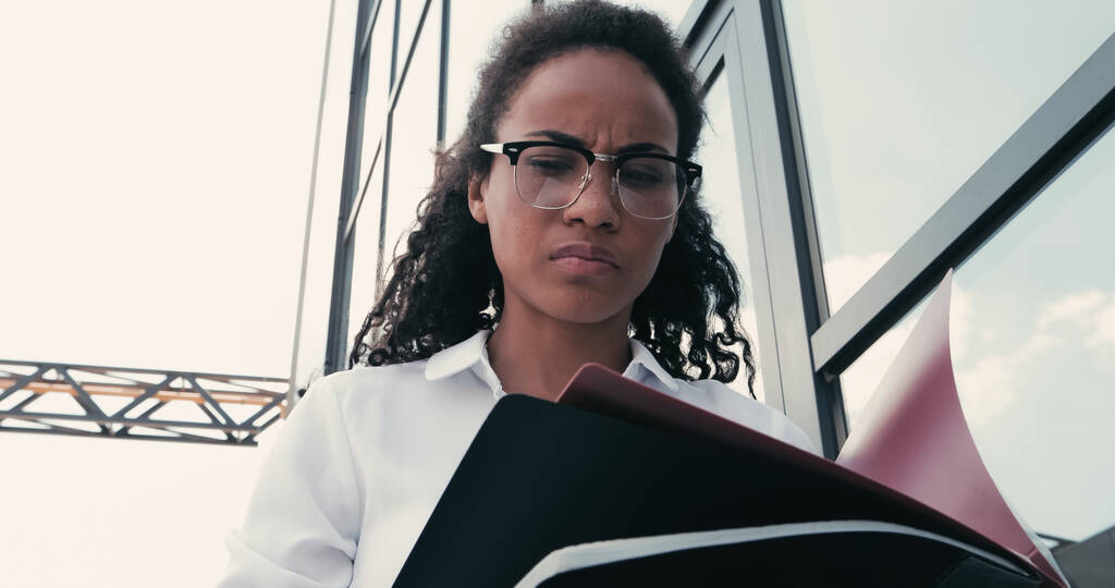 laag hoek uitzicht van krullend Afrikaans Amerikaans zakenvrouw in bril kijken naar papieren mappen in de buurt van gebouw  - Foto, afbeelding