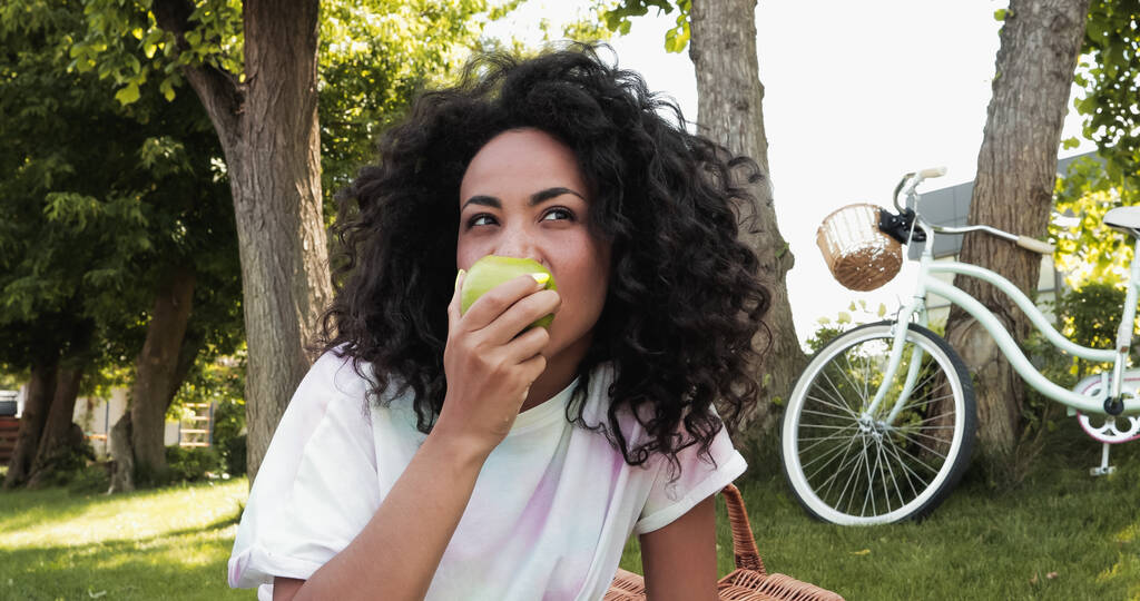 mujer afroamericana mordiendo manzana fresca cerca de bicicleta en el parque  - Foto, imagen