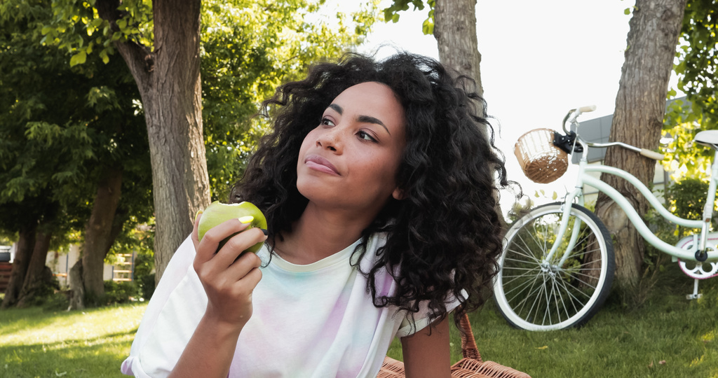 curly african american woman eating fresh apple near bicycle in park  - Photo, Image