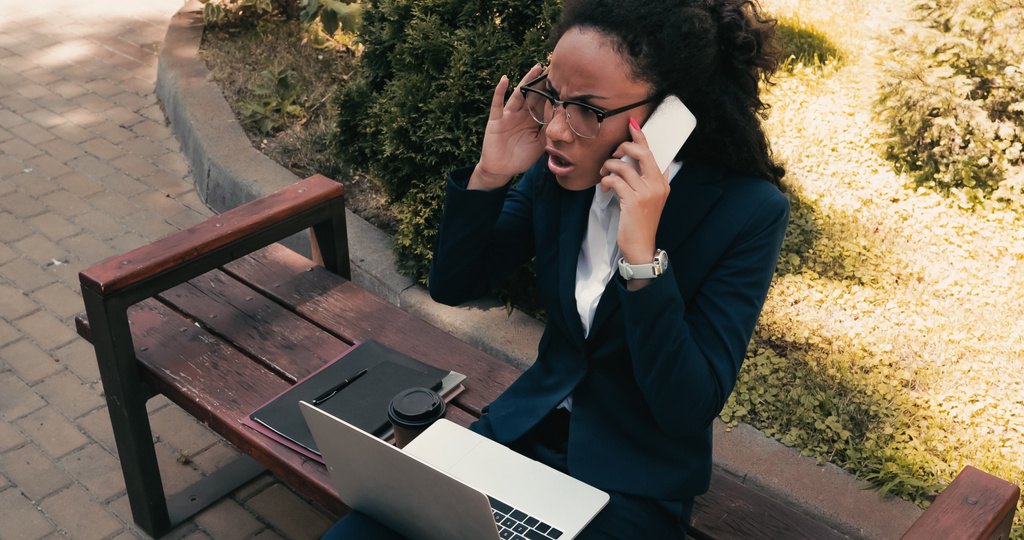 elégedetlen afro-amerikai üzletasszony beszél okostelefonon, miközben ül padon laptoppal  - Fotó, kép