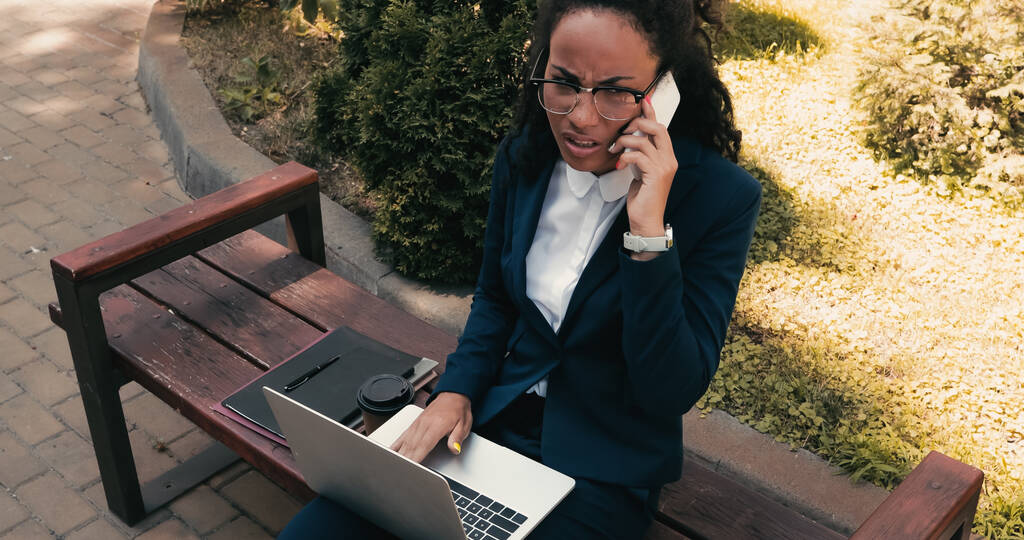 frustrada mujer de negocios afroamericana hablando por teléfono celular mientras se sienta en el banco con el ordenador portátil  - Foto, imagen
