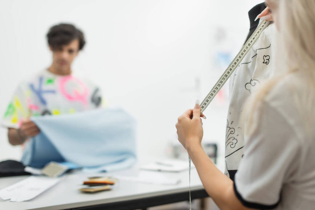 diseñador de moda medir ropa en maniquí cerca borrosa colega sosteniendo muestra de tejido - Foto, imagen