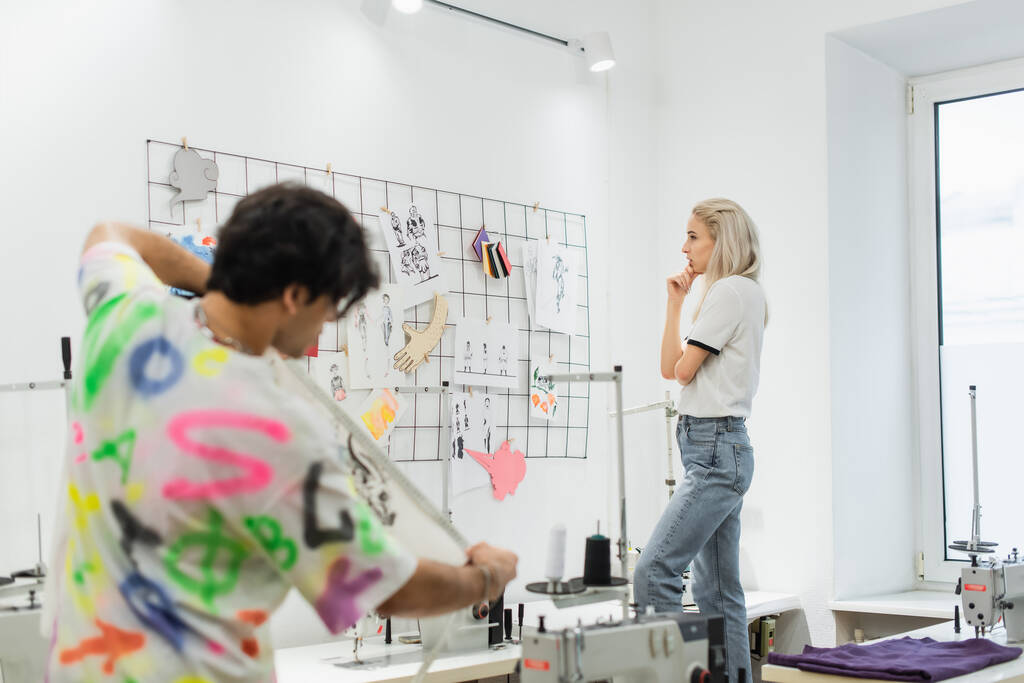 Blonde Modedesignerin denkt an Skizzen an der Wand und verschwommene Kollegin mit Maßband - Foto, Bild