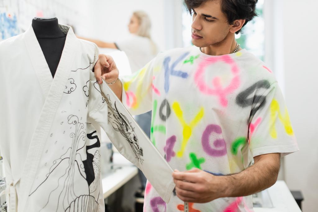 Moda tasarımcısı, bulanık bir arkaplan üzerinde çalışan manken ve meslektaşının kimonosuna bakıyor. - Fotoğraf, Görsel