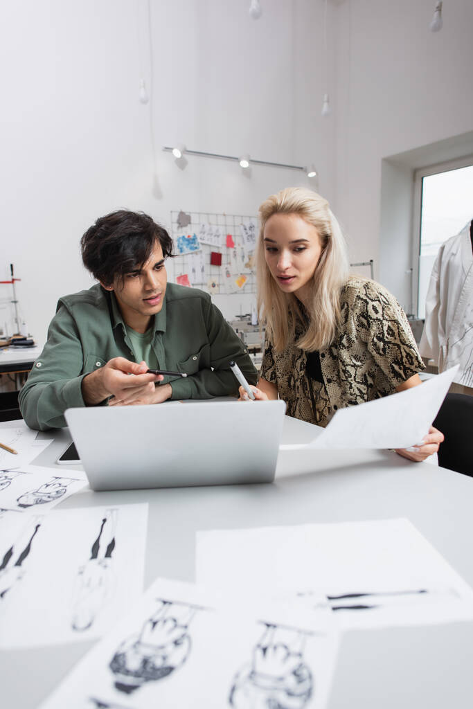 молодий дизайнер вказує на ноутбук біля блондинки колеги і розмиті ескізи
 - Фото, зображення