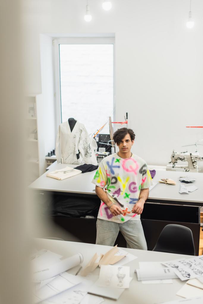 vista de ángulo alto del diseñador de moda de moda mirando a la cámara cerca del escritorio con patrones de costura, primer plano borroso - Foto, Imagen