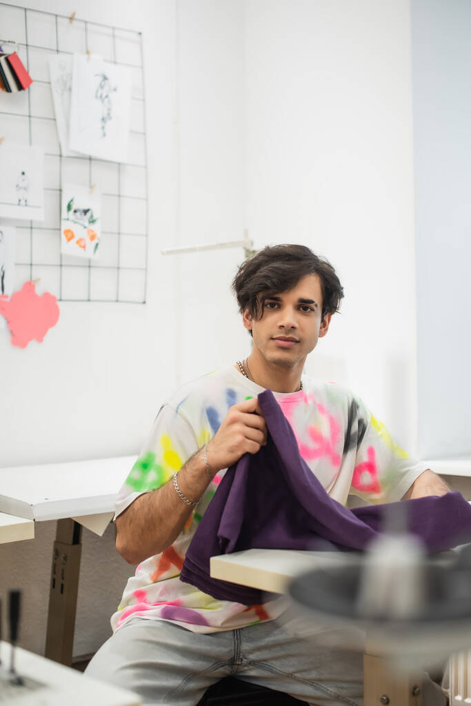 jovem designer olhando para a câmera enquanto segurando amostra de tecido no atelier de moda - Foto, Imagem