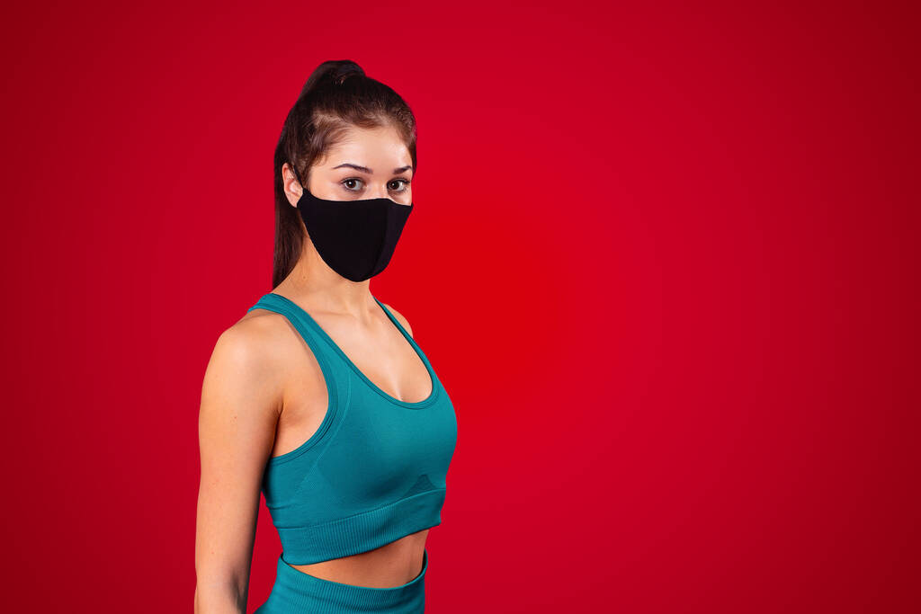 Спортивная молодая женщина позирует в черной многофункциональной маске, одетая в спортивную форму на красном фоне. Высокое качество фото - Фото, изображение