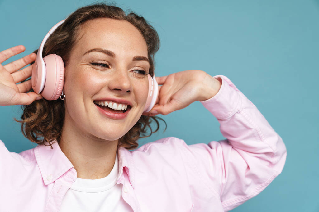 Jeune femme rousse riant en écoutant de la musique avec des écouteurs isolés sur fond bleu - Photo, image