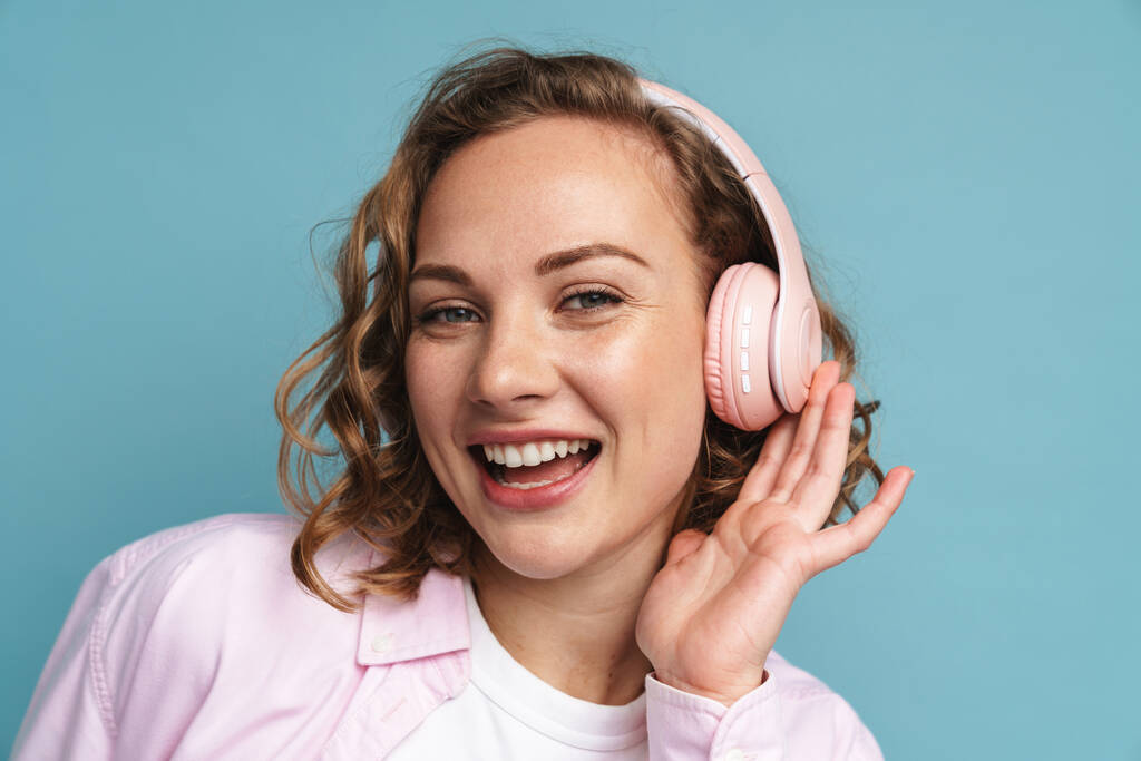 Genç kızıl kadın, mavi arka planda izole edilmiş kulaklıklarla müzik dinlerken gülüyor. - Fotoğraf, Görsel