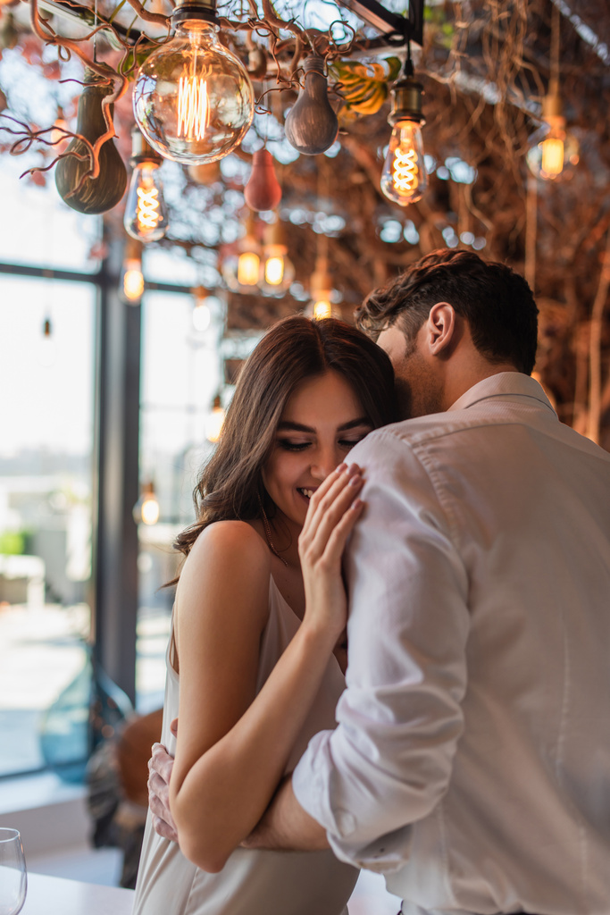 άντρας να φιλάει και να αγκαλιάζει χαρούμενη νεαρή γυναίκα στο εστιατόριο  - Φωτογραφία, εικόνα