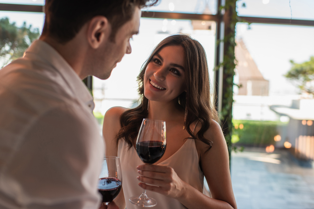 fröhliche Frau hält Glas Wein in der Hand und blickt verschwommenen Mann an  - Foto, Bild