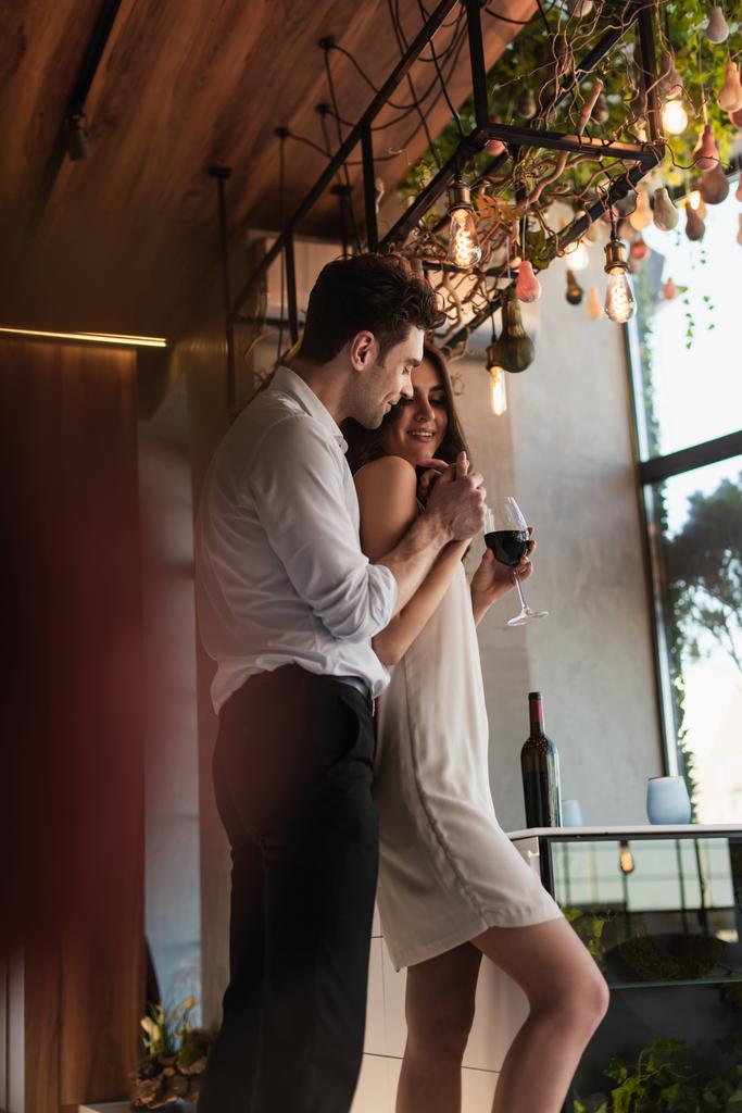 homme joyeux étreignant petite amie heureuse en robe glissante avec un verre de vin - Photo, image