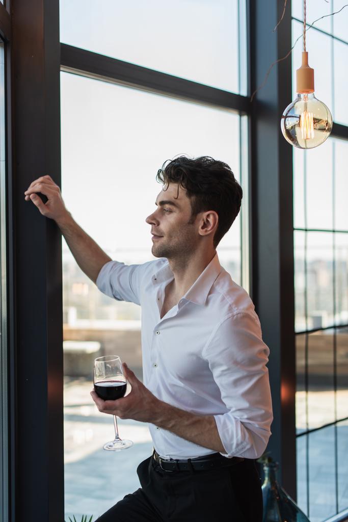 窓の近くに立っている間に赤ワインのグラスを持っている白いシャツのエレガントな男 - 写真・画像