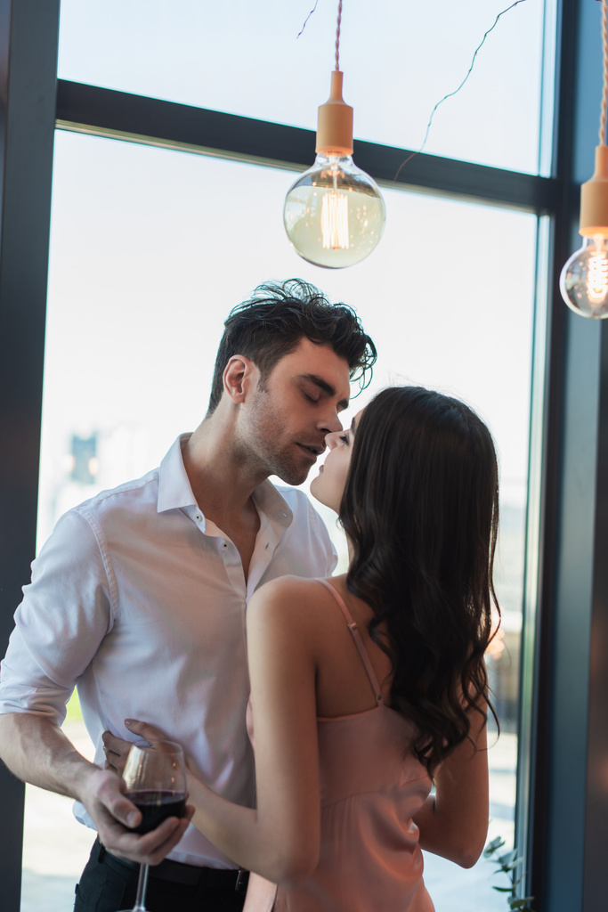 Elinde kırmızı şarap bardağı tutan ve slip elbiseli esmer kadını öpen bir adam. - Fotoğraf, Görsel