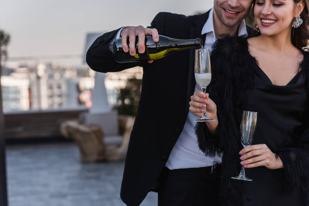 happy man pouring champagne in glass of girlfriend in faux fur jacket  - Photo, Image