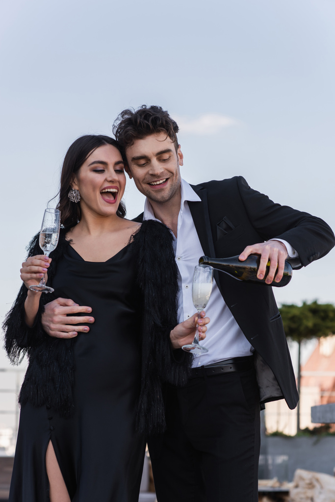 happy man pouring champagne in glass of excited girlfriend in faux fur jacket  - Photo, Image