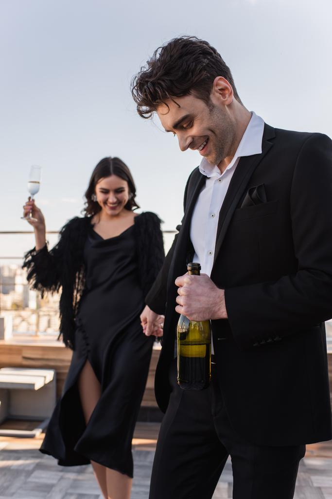 happy man with bottle of champagne holding hand of blurred and cheerful girlfriend  - Photo, Image