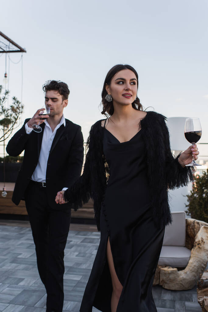 mujer feliz cogida de la mano con el hombre beber vino mientras camina en la terraza  - Foto, Imagen