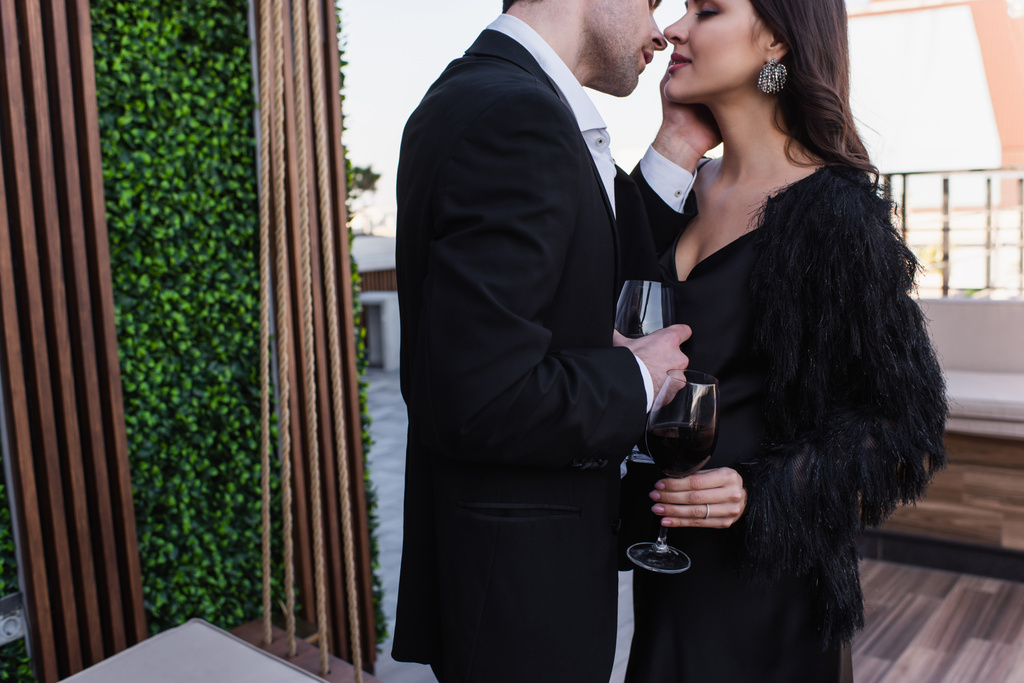 homem beijando mulher e segurando copo de vinho tinto no terraço  - Foto, Imagem
