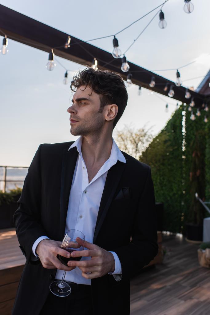 elegantní muž v obleku drží sklenici červeného vína a hledá cestu na terase  - Fotografie, Obrázek