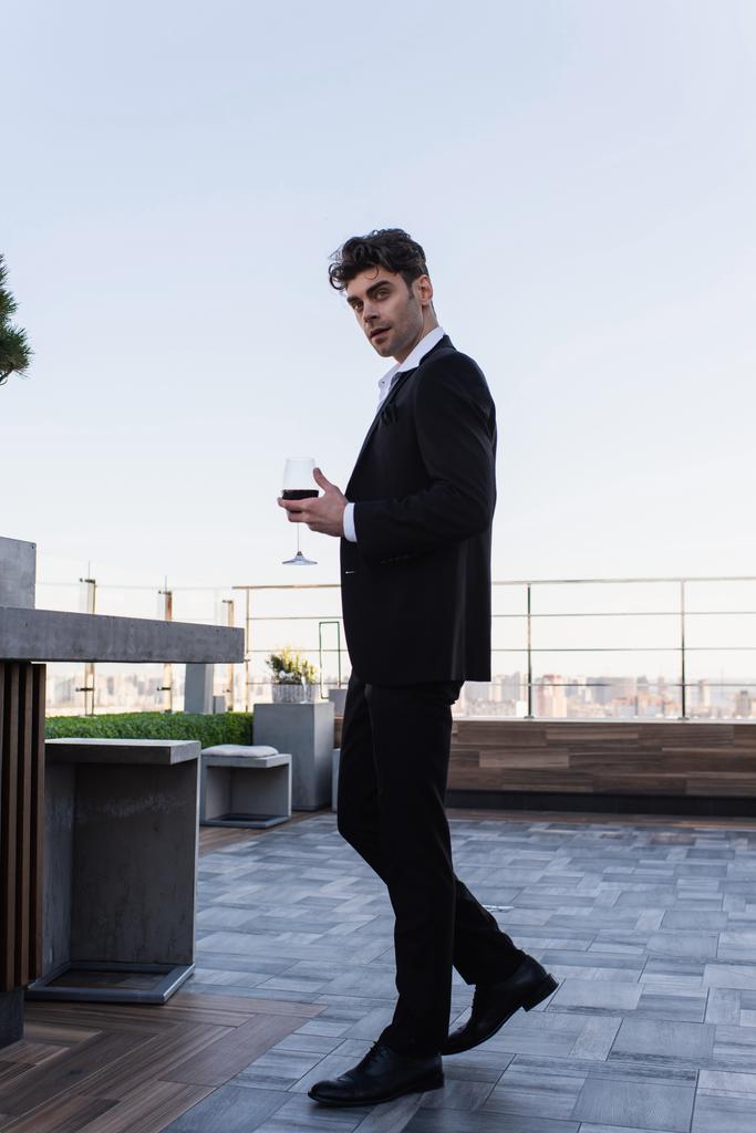 full length of elegant man in suit holding glass of red wine on terrace  - Photo, Image