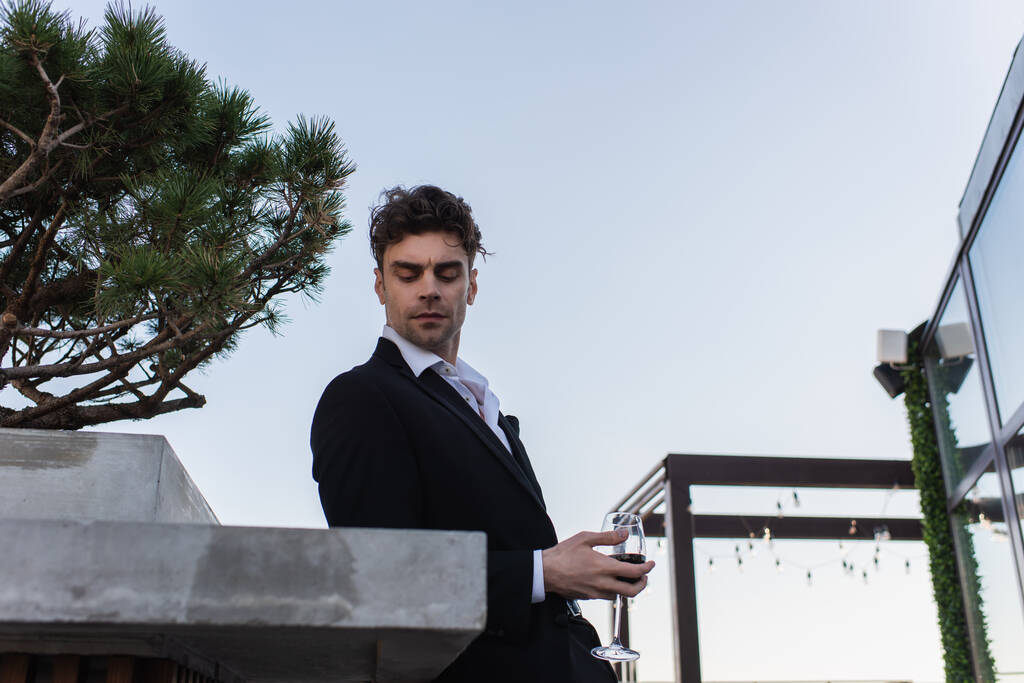hombre guapo en traje sosteniendo copa de vino tinto en la terraza  - Foto, Imagen