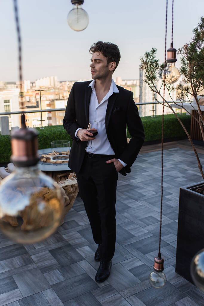 повна довжина елегантного чоловіка в костюмі, що стоїть з рукою в кишені і тримає келих червоного вина на терасі
  - Фото, зображення