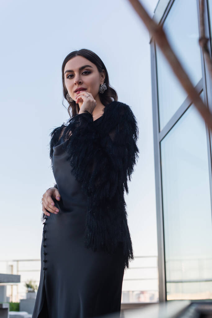 lage hoek uitzicht op jonge vrouw in zwarte slip jurk en faux fur jas op het terras  - Foto, afbeelding