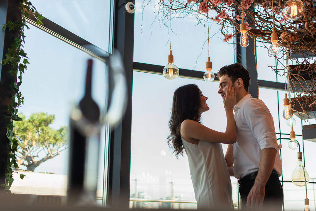 πλαϊνή άποψη του ευτυχισμένη γυναίκα σε ολίσθηση φόρεμα αγγίζοντας το πρόσωπο του φίλου στο εστιατόριο  - Φωτογραφία, εικόνα