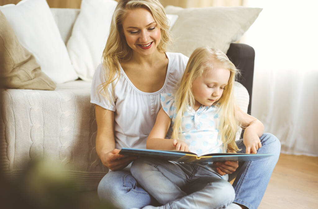 Mutlu bir aile. Sarışın genç anne ahşap zeminde otururken sevimli kızına kitap okuyor. Annelik kavramı - Fotoğraf, Görsel