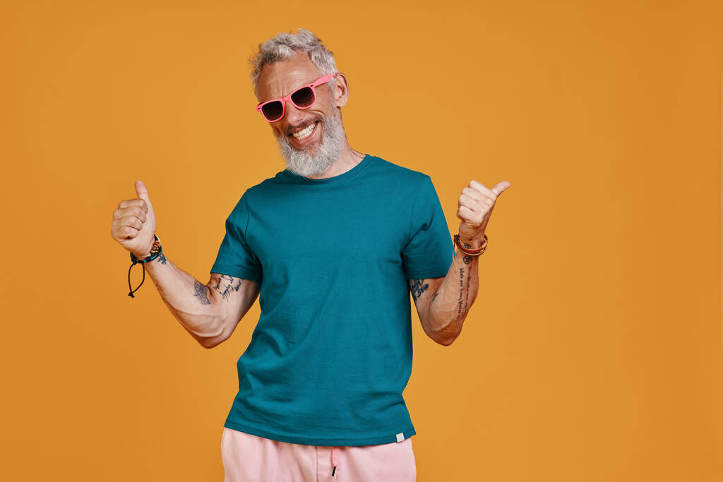 Stylish senior man gesturing and smiling while standing against orange background - Photo, Image