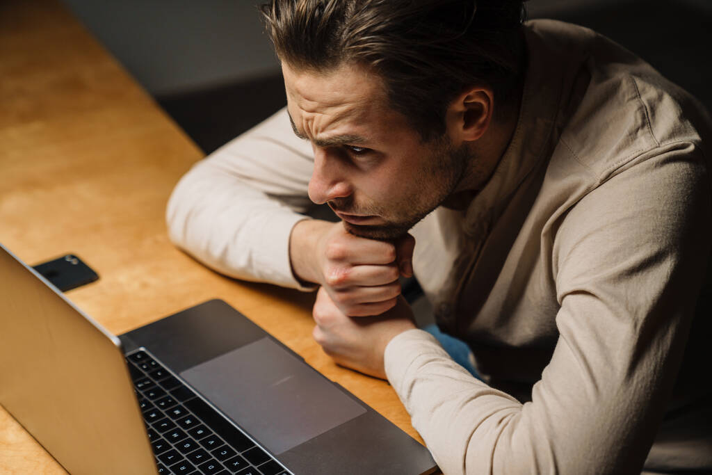Усталый брюнетка среднего возраста мужчина смотрит на экран ноутбука, сидя в офисе - Фото, изображение