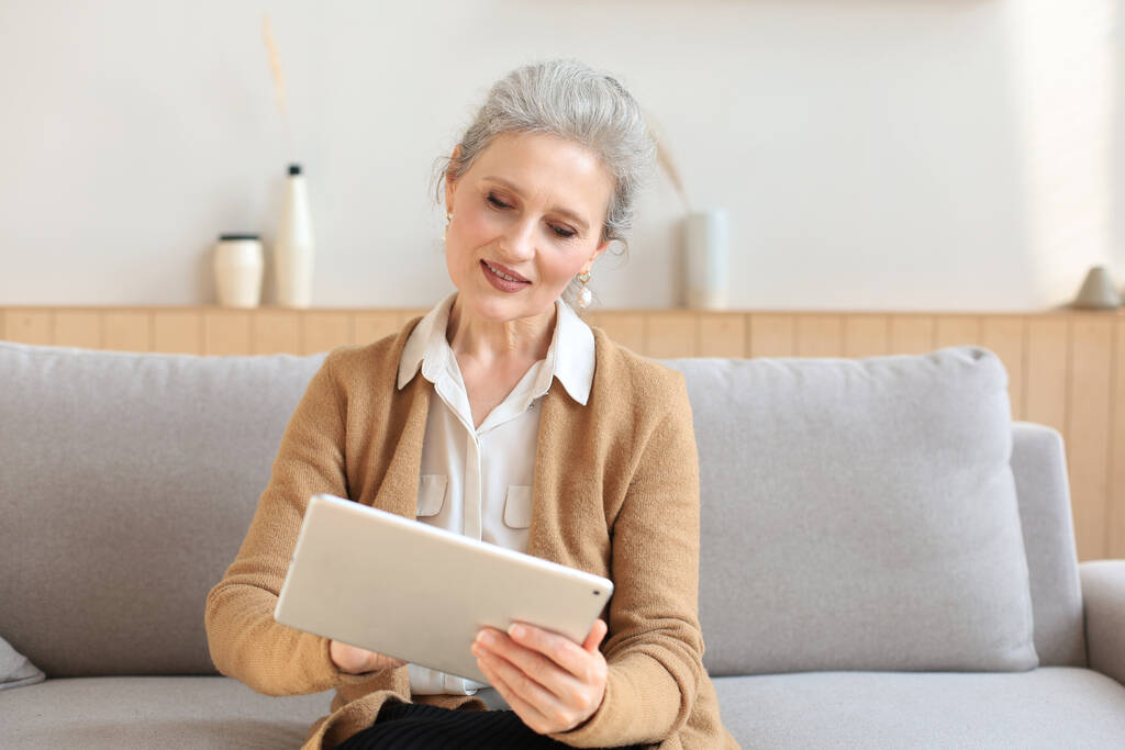 Vidám középkorú nő ül a kanapén, számítógépes tablet alkalmazások, nézi a képernyőn, olvasás jó hír a szociális hálón, vásárlás vagy csevegés online - Fotó, kép