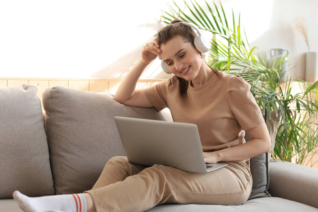 Giovane donna sorridente con cuffie e laptop sul divano - Foto, immagini