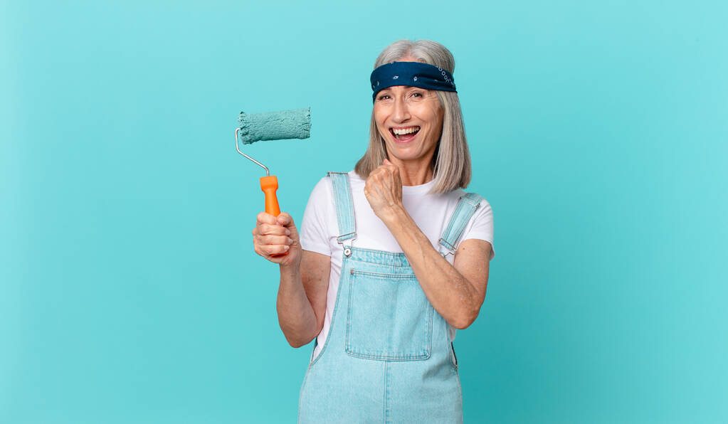 mujer de pelo blanco de mediana edad sintiéndose feliz y enfrentándose a un desafío o celebrando con un rodillo pintando una pared - Foto, imagen