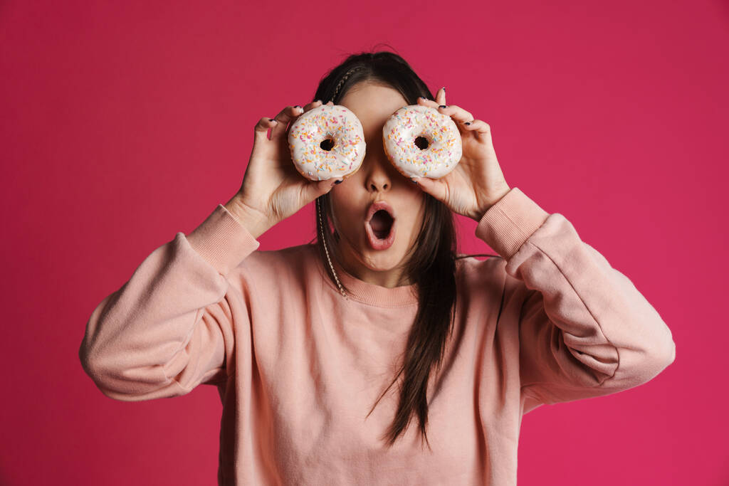 Ευτυχισμένη νεαρή γυναίκα που ποζάρει κρατώντας ντόνατς πάνω από ροζ φόντο στον τοίχο - Φωτογραφία, εικόνα